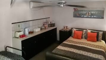 A compact hotel room with a bed, a small kitchenette, a ceiling fan, and a painting on the wall.