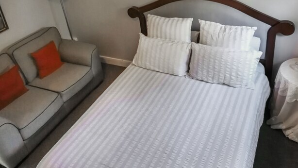 Una cama bien hecha con ropa de cama blanca y varios almohadones, un sofá gris con un cojín rojo, y una lámpara sobre una mesa.