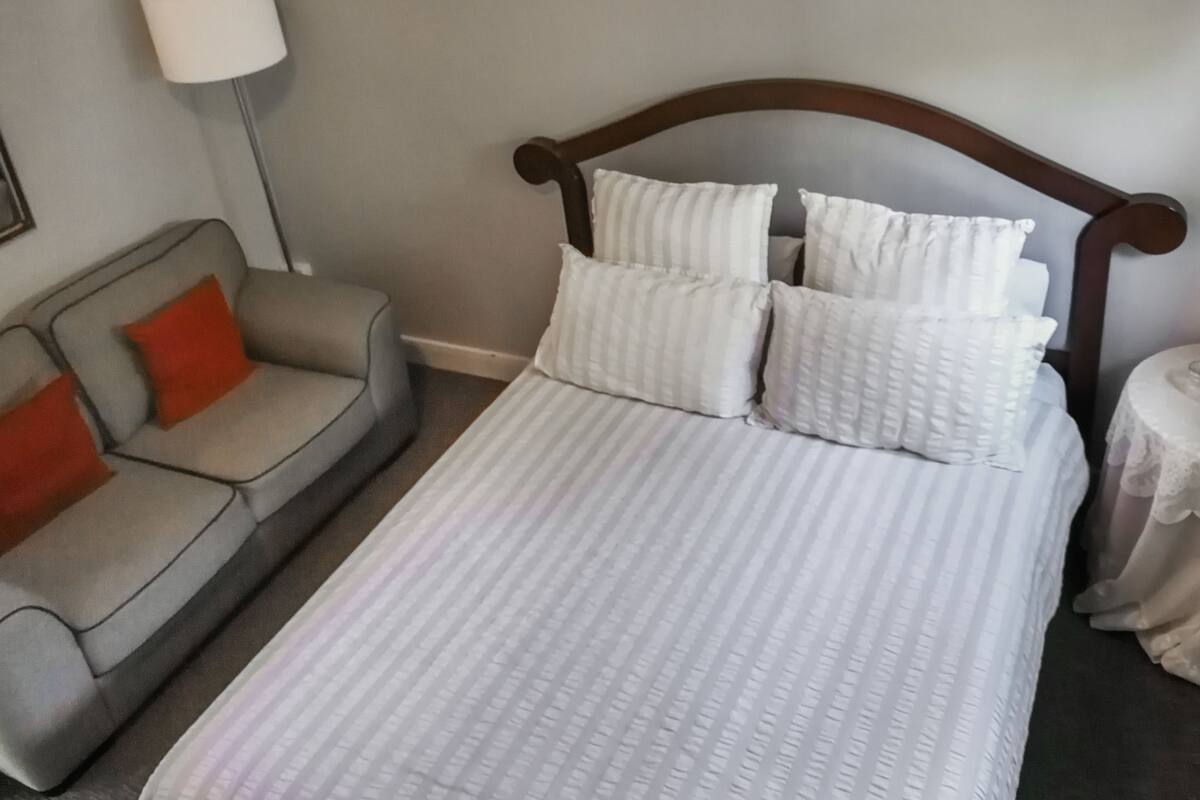 Una cama bien hecha con ropa de cama blanca y varios almohadones, un sofá gris con un cojín rojo, y una lámpara sobre una mesa.