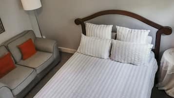 A neatly made bed with white bedding and multiple pillows, a grey sofa with a red cushion, and a lamp on a table.