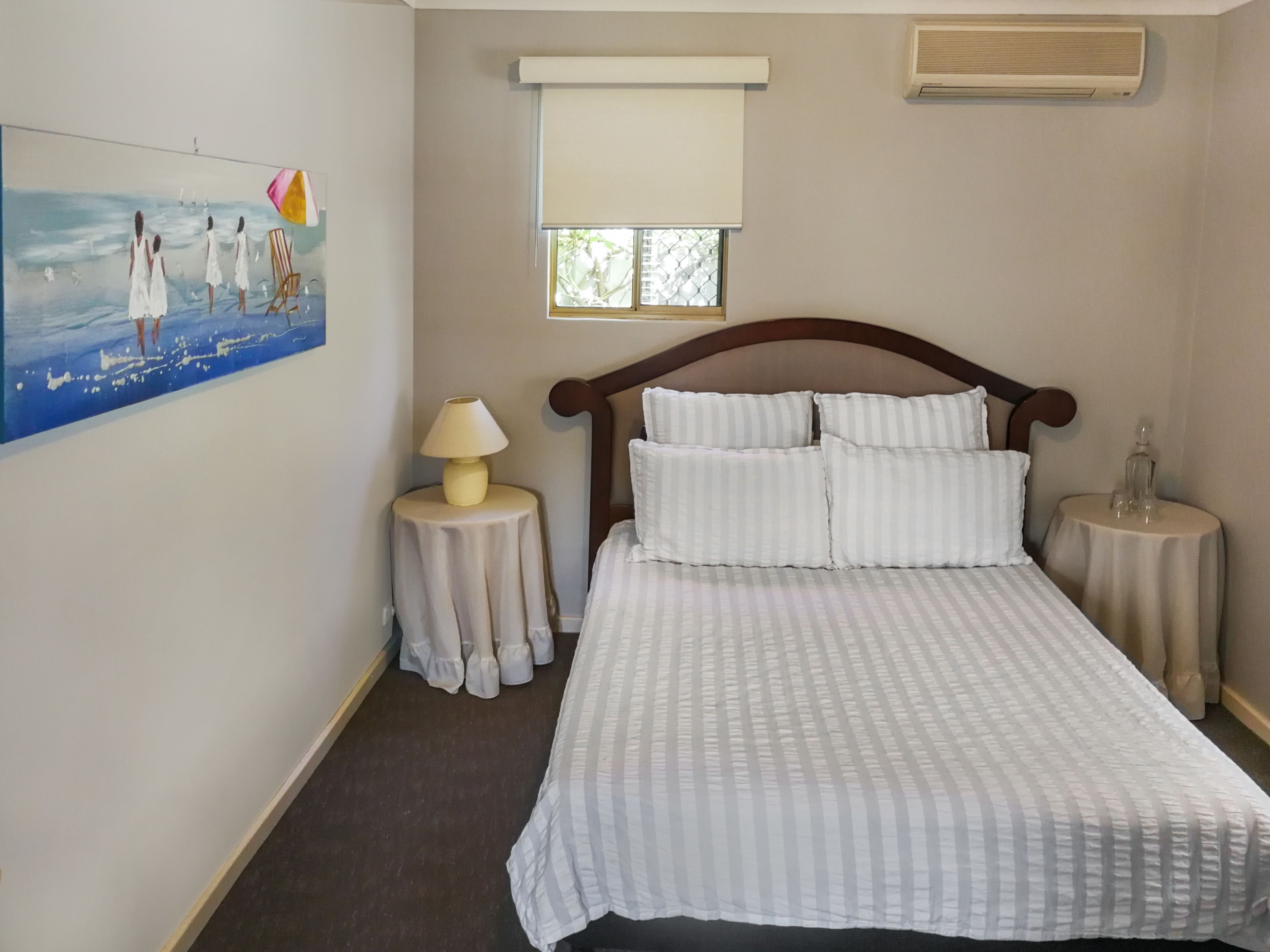 Un dormitorio con cama, mesita de noche, lámpara, ventana con persianas y un cuadro en la pared.