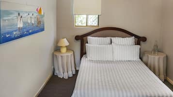 Un dormitorio con cama, mesita de noche, lámpara, ventana con persianas y un cuadro en la pared.
