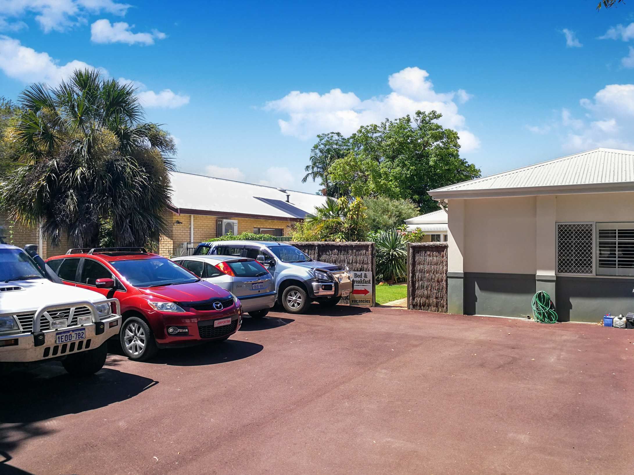 Un estacionamiento con varios coches, un edificio con un cartel que indica servicios de casa de huéspedes, y una zona ajardinada cercada.