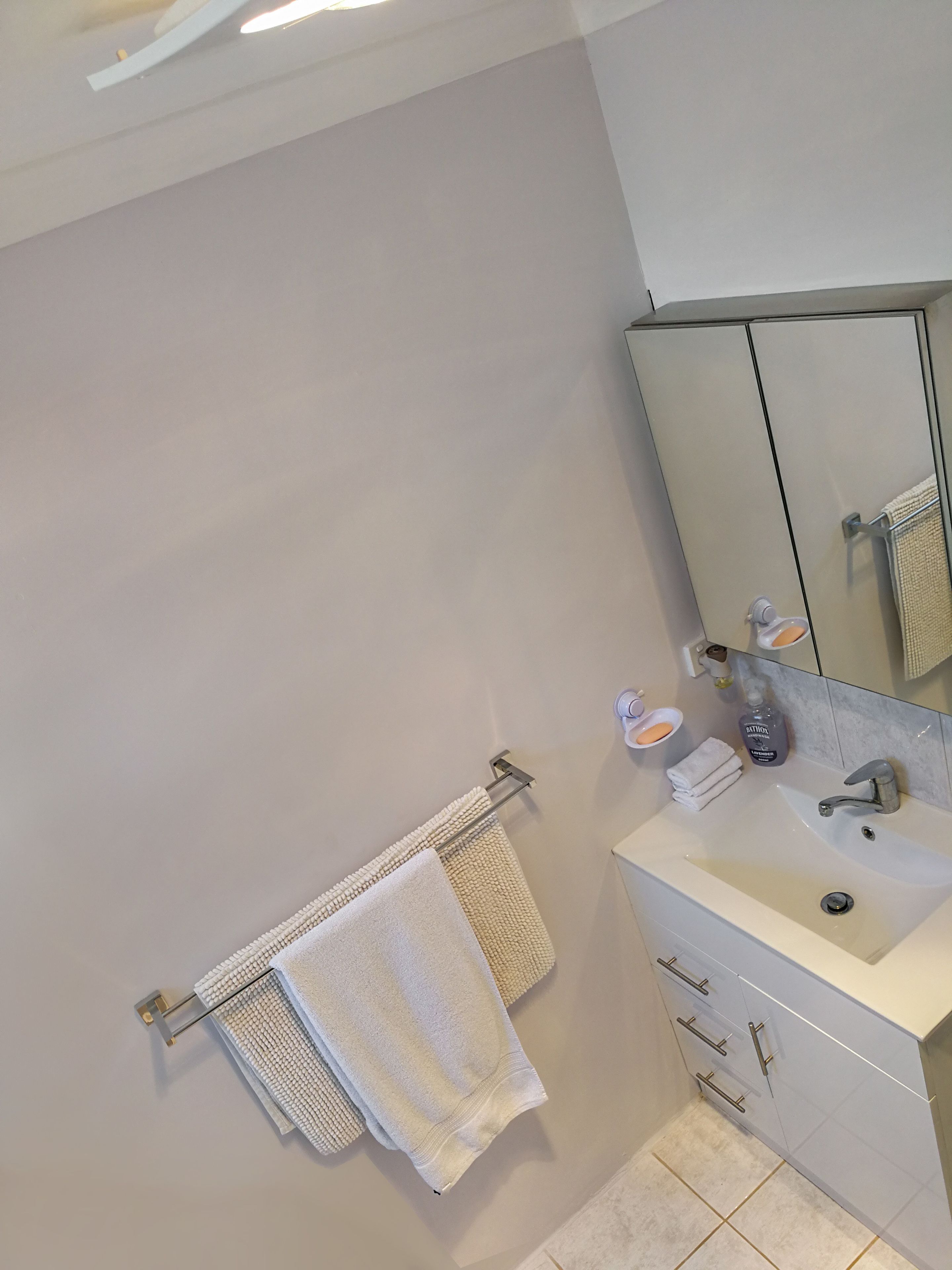 A modern bathroom with a white sink, mirror, and cabinets. There are two towels hanging on hooks and a bottle of hand soap on the counter.