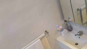 A modern bathroom with a white sink, mirror, and cabinets. There are two towels hanging on hooks and a bottle of hand soap on the counter.