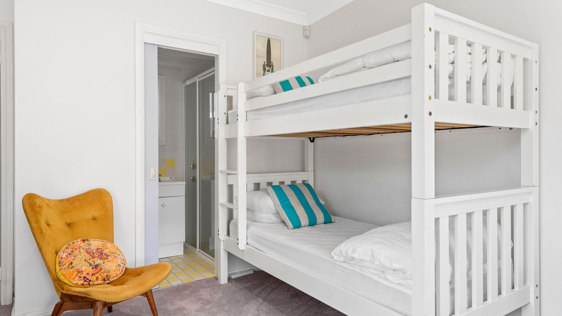 A bunk bed with white bedding and a striped pillow, a mustard-colored chair with a floral cushion, and a doorway leading to a bathroom.