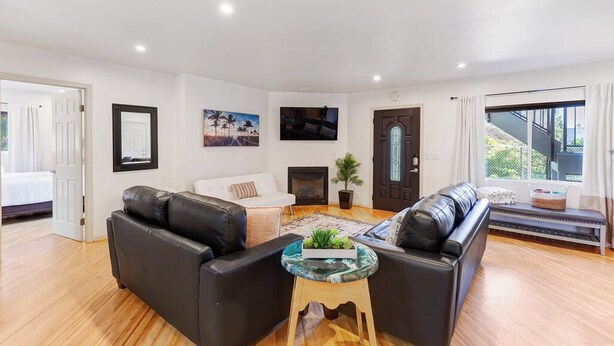 A modern living room with a black leather sofa, a white sofa, and a glass coffee table with plants. There is a fireplace, a door with a mirror, and a large window with curtains. - Point Loma 3 bedroom/2bath (San Diego)