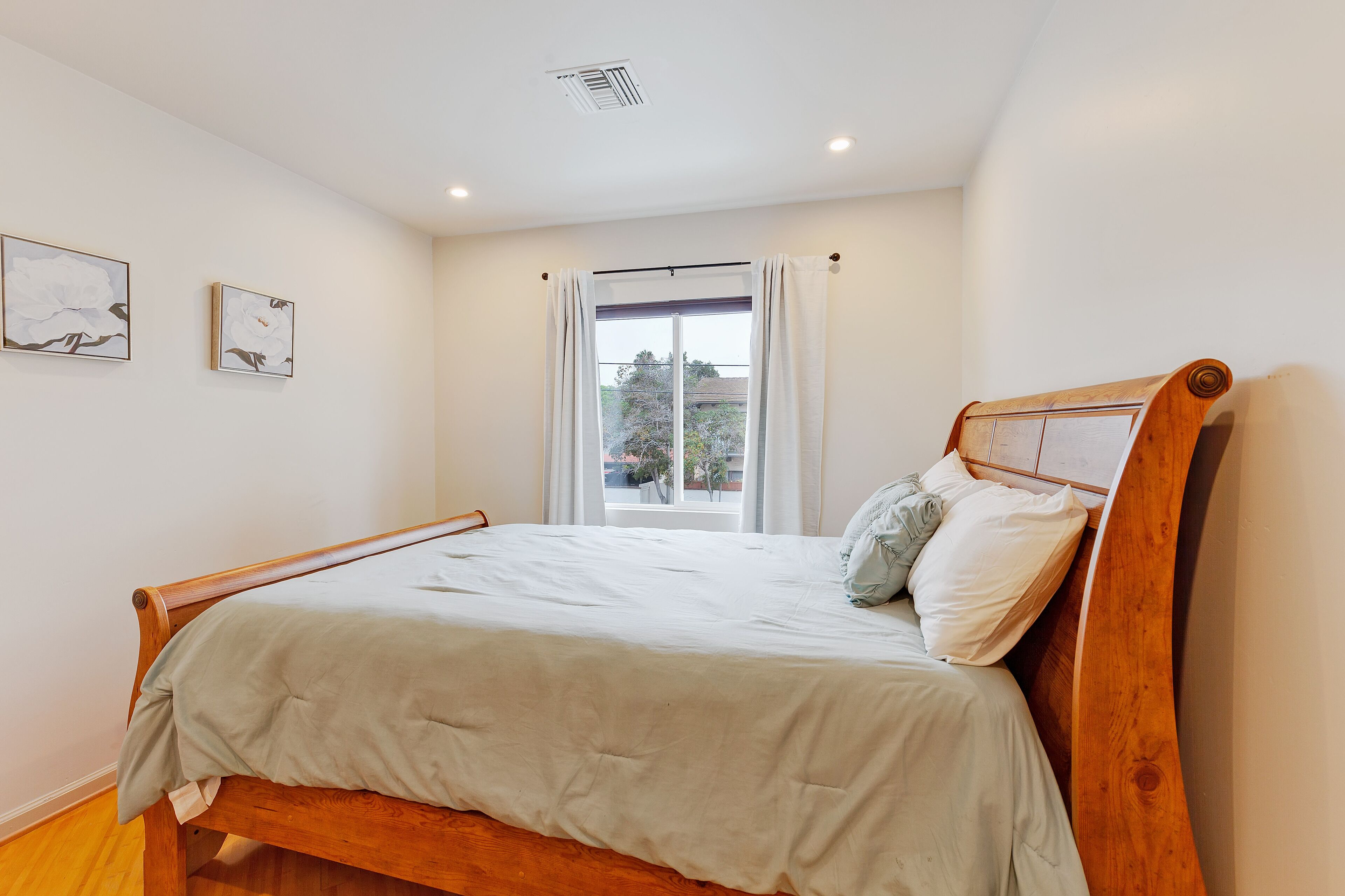 Un dormitorio con una cama de madera, ropa de cama blanca, un ventanal con cortinas y dos obras de arte colgadas en la pared.