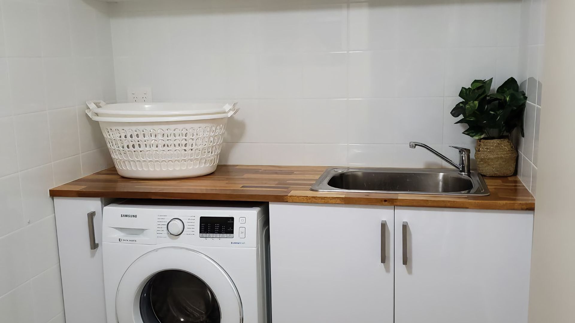 Un espacio de lavandería pequeño con lavadora, un lavabo y una cesta blanca sobre una encimera de madera.
