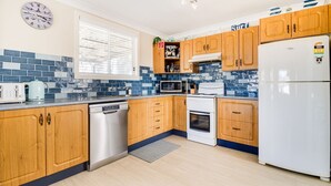 A kitchen with wooden cabinets, a white refrigerator, a microwave, a dishwasher, and a clock on the wall. - The Perfect Holiday Home (Sussex Inlet)