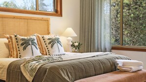 Una cama bien hecha con cabecera, almohadas con estampado de hojas de palma, una lámpara de noche y una ventana con cortinas.