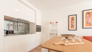Una cuina moderna amb armaris blancs, una taula de dinar de fusta i una cadira blanca. Hi ha obres d'art emmarcades a la paret i una estanteria blanca amb objectes decoratius.
