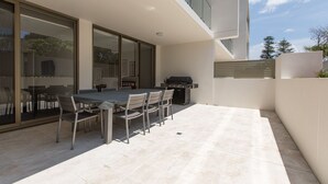 Un pati exterior modern amb taula i caires de dinar, una barbacoa i una porta de vidre que dona a una zona coberta.