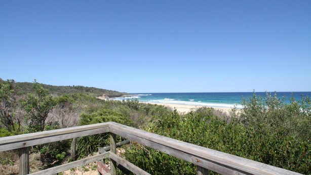 Une promenade en bois mène à une plage d’où l’on a une vue dégagée sur l’océan, avec un paysage vallonné en arrière-plan.