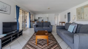 A modern living room with a sofa, a coffee table, a TV, and a kitchen in the background. - Holiday on the Water (Sussex Inlet)