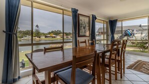 Un espace repas comprenant une table et des chaises, de grandes fenêtres avec des rideaux, et une vue sur un lac et des bâtiments.
