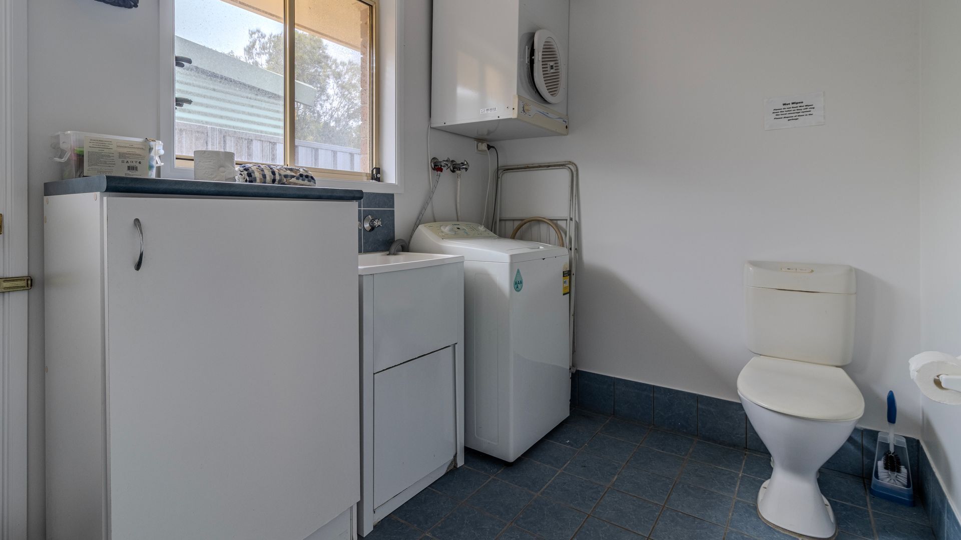 A compact bathroom with a toilet, a washing machine, a dryer, and a window with a view of a house.