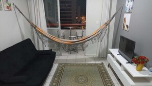 Una sala de estar con un sofá negro, un televisor sobre un mueble blanco, un tapete estampado verde y una hamaca.