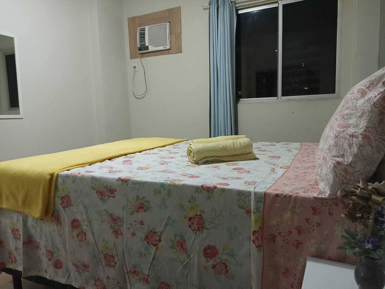 A room with a bed covered in floral bedding, a window with curtains, and a wall-mounted air conditioner.