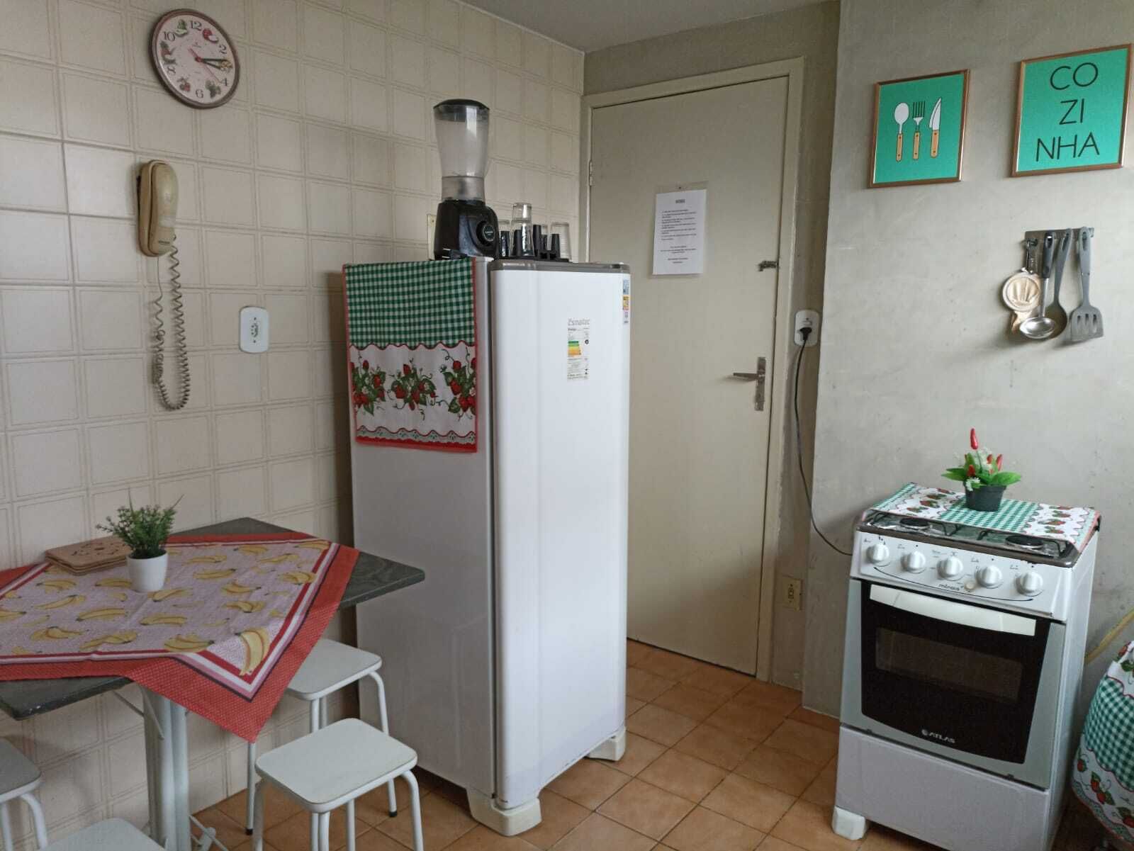 A compact kitchen with a refrigerator, a stove, a small table with a banana-patterned tablecloth, and a single chair.