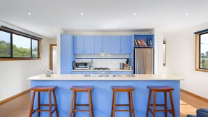 A modern kitchen with blue cabinets, a central island with stools, stainless steel appliances, and a view of the outdoors through large windows.