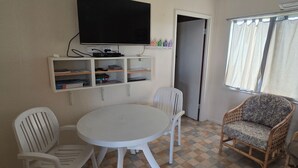 Una habitació amb una taula rodona blanca, dues cadires blanques, una cadira de vimet i una televisió penjada a la paret, a més d'una estant amb llibres i aparells electrònics.