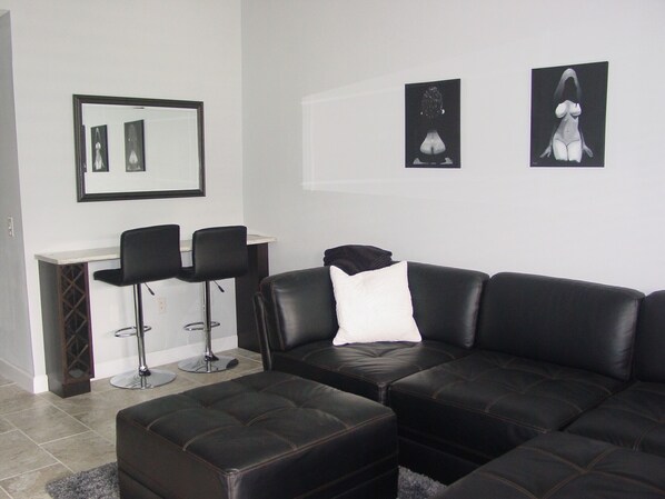 Un salon moderne avec un canapé d’angle en cuir noir, deux tabourets de bar noirs, une petite table blanche et un miroir encadré accroché au mur.