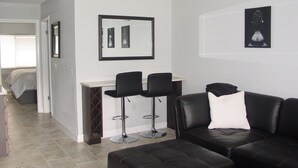A modern living room with a black leather sofa, a coffee table, and two black bar stools. There is a bed with a white headboard in the background, and a framed picture on the wall.
