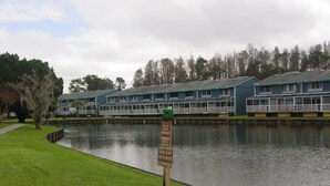 A lakeside view with a sign warning against feeding or molesting alligators, a wooden sculpture, and a row of blue buildings with a grassy area in the foreground.