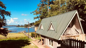 A wooden cabin with a metal roof, situated by a body of water with boats and docks in the background. - Privacy, fishing, pool--Just 10 mins to beach/base (Sneads Ferry)