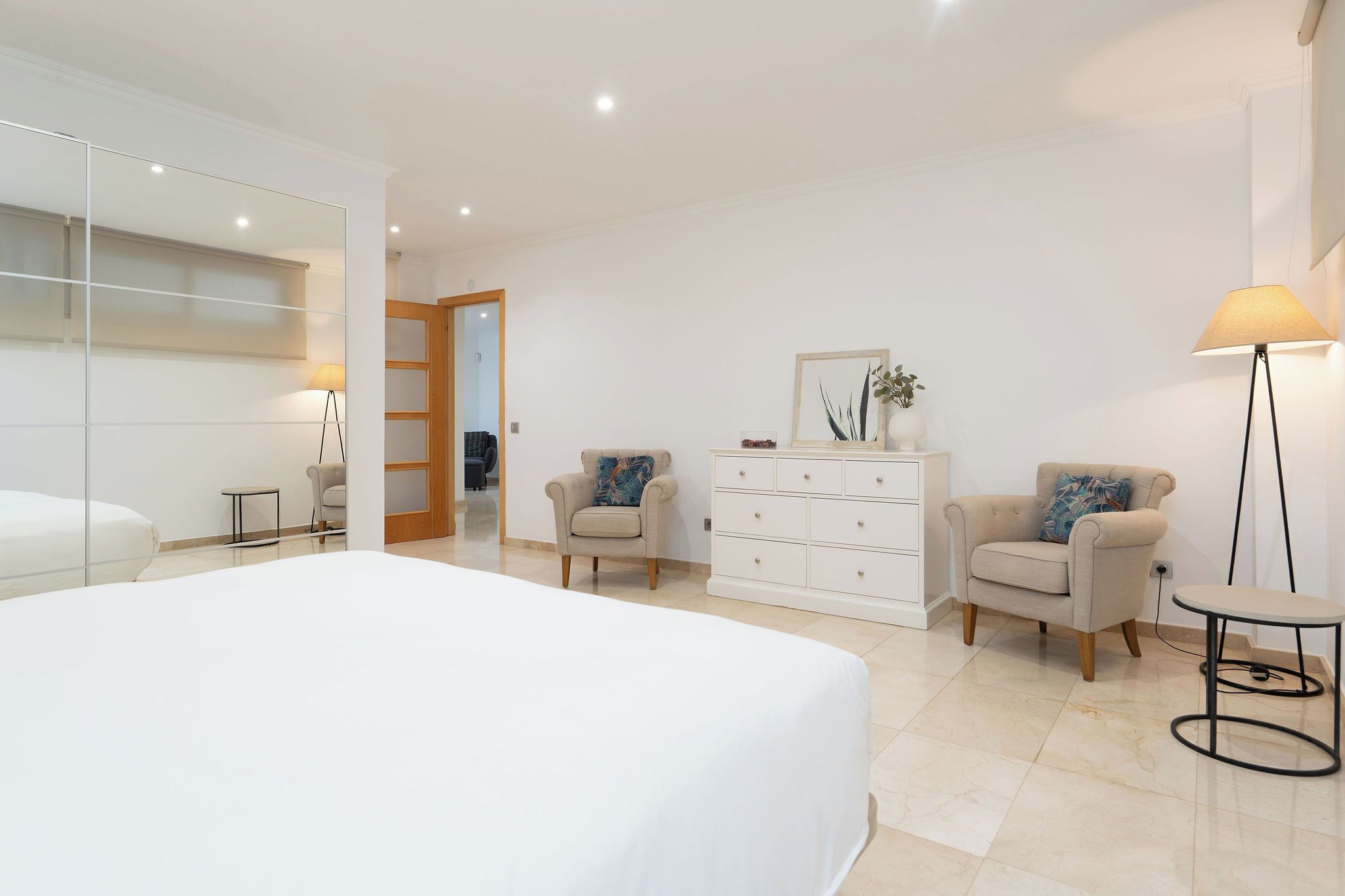 Un dormitorio moderno con una cama grande, un aparador, dos sillones, una mesita y una lámpara.
