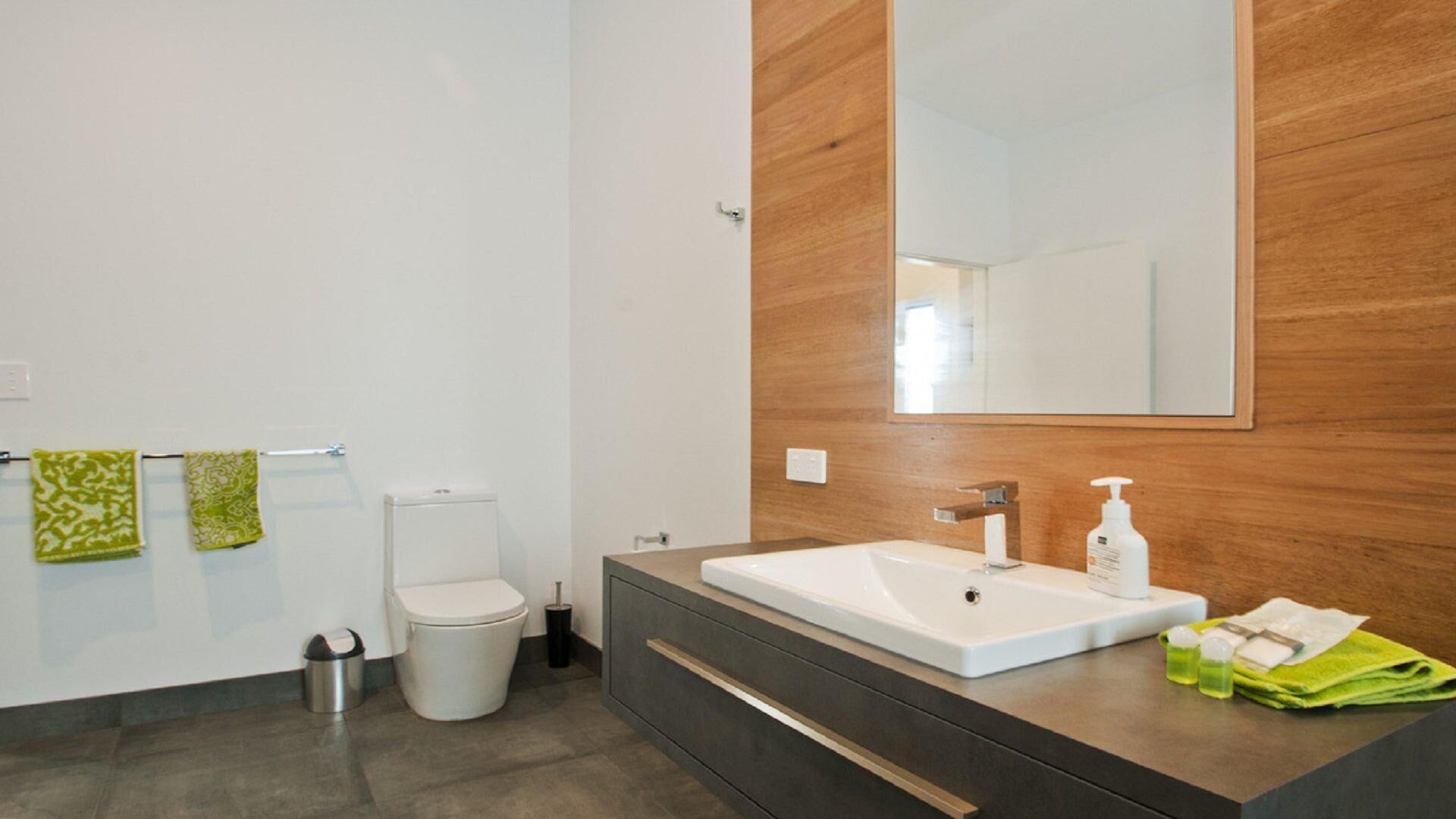 Un baño moderno con inodoro blanco, tocador de madera con lavamanos y un espejo grande.