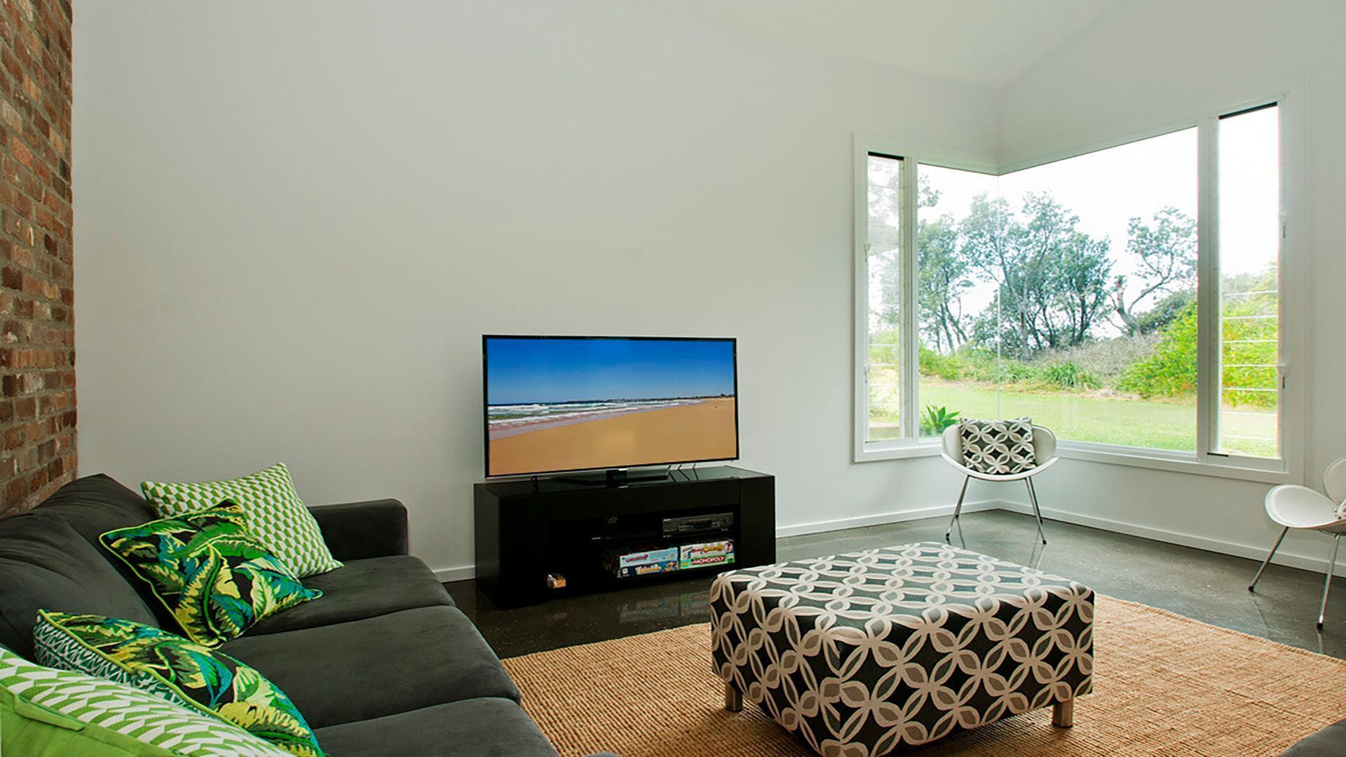 Un salon moderne avec un téléviseur à écran plat posé sur un meuble noir, un canapé gris agrémenté de coussins à motifs, un pouf à motifs et une grande fenêtre donnant sur un paysage verdoyant.