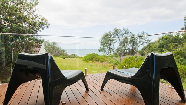 Two black plastic chairs on a wooden deck with a glass balustrade, overlooking a grassy area and the sea. - Farrant House - direct access to beach (Culburra Beach)