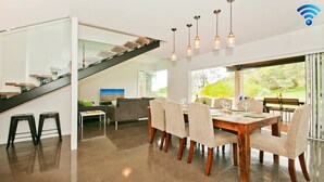 A modern dining area with a wooden table and chairs, a staircase, and a view of greenery outside. - Farrant House - direct access to beach (Culburra Beach)