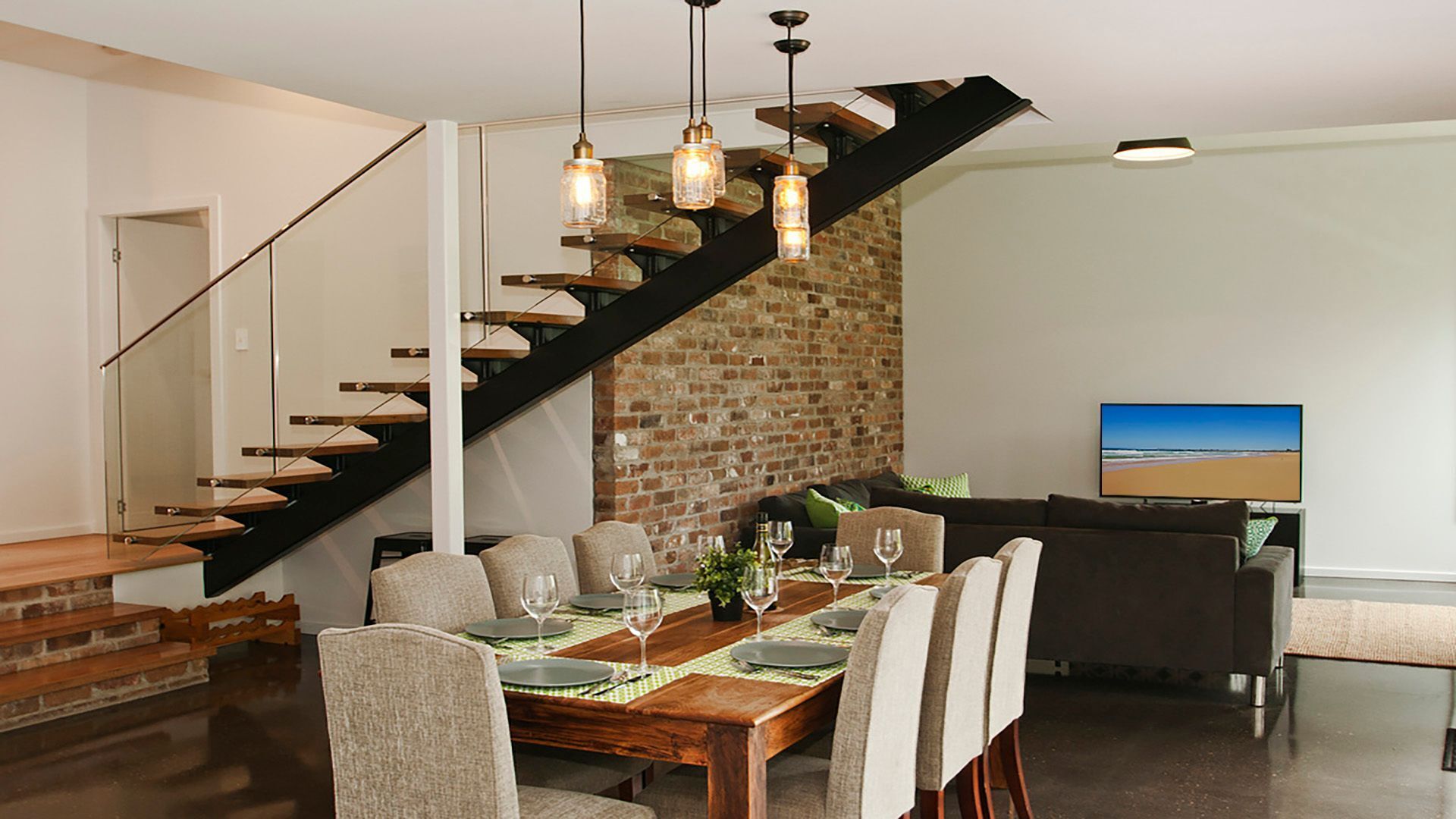 A modern dining area with a wooden table set for six, a brick accent wall, a staircase, and a flat-screen TV displaying a beach scene.