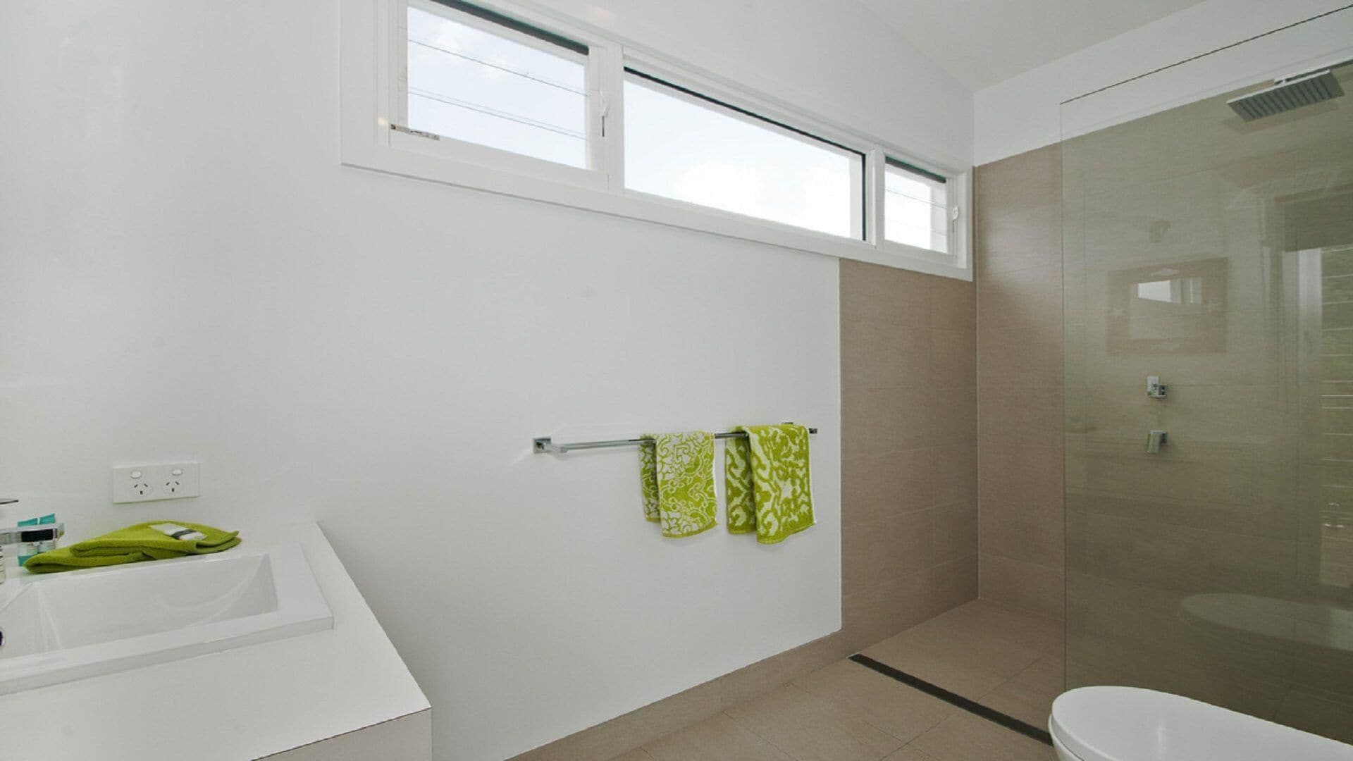 Ein modernes Badezimmer mit Dusche, Waschbecken und Toilette. Zwei Fenster sorgen für natürliches Licht, und ein Handtuchhalter trägt zwei Handtücher.