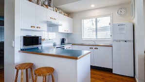 Een moderne keuken met witte kasten, een ingebouwde magnetron, een spoelbak en een koelkast. Er staan twee houten krukken en er is een raam met zonwering.