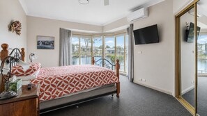 A bedroom with a bed, a desk, a chair, a TV, and a view of a body of water. - Cormorant Waterfront Home (Sussex Inlet)