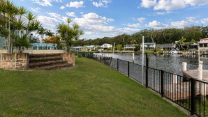 A waterfront property with a grassy lawn, a dock, and a row of houses in the background. - Cormorant Waterfront Home (Sussex Inlet)