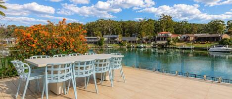 A lakeside patio with a dining table and chairs, surrounded by a swimming pool and residential buildings.