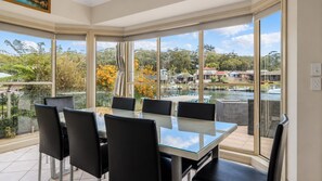 A dining area with a glass table and chairs, large windows, and a view of a body of water and houses.