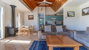 A living room with a sofa, a wooden coffee table, and a fireplace. There is a kitchen in the background with teal cabinets and a hanging light fixture. - Sea Shell Chalet @ Berrara Chalet (Berrara)