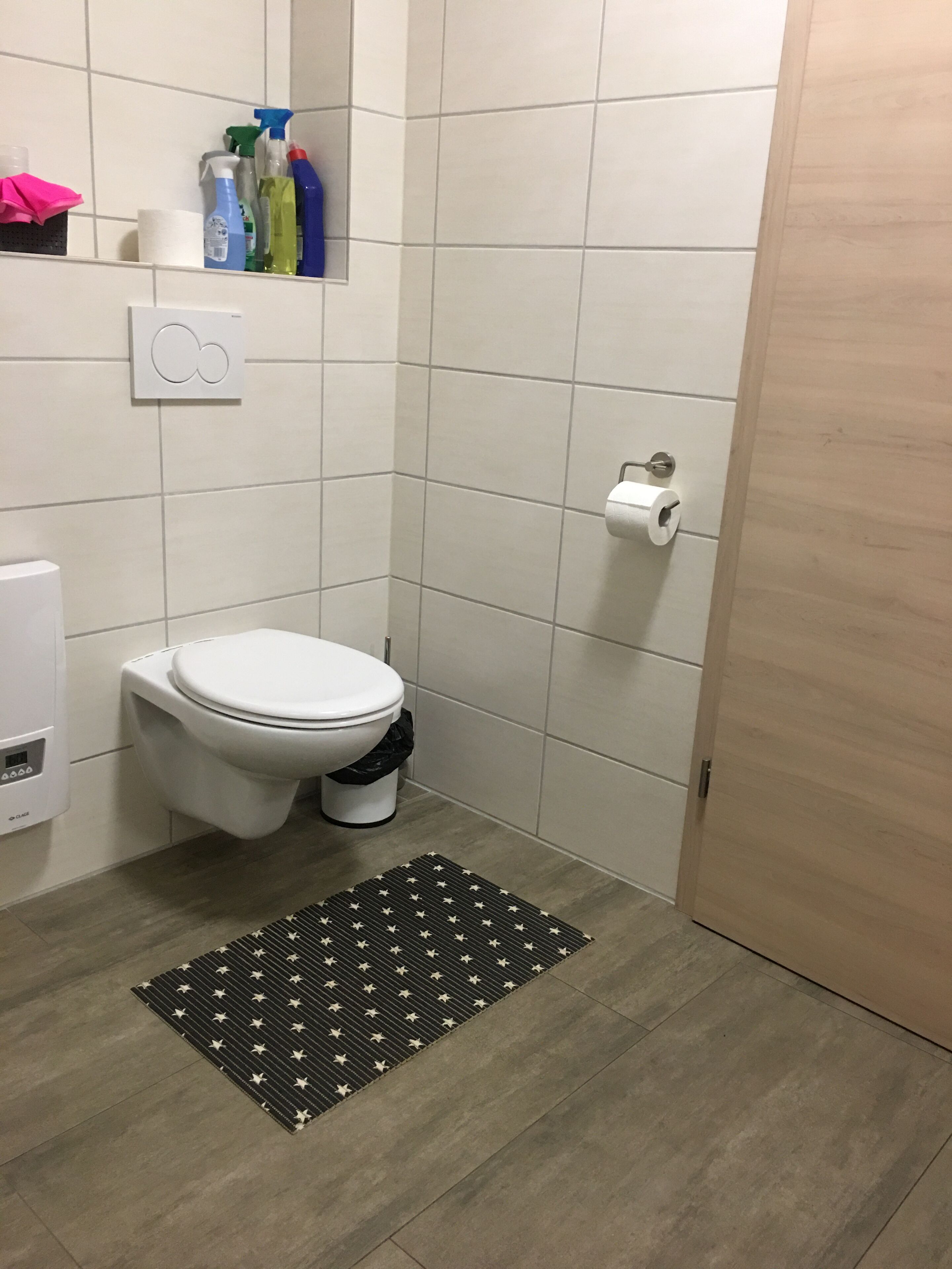 Ein kleines Badezimmer mit Toilette, einem Händetrockner, einem Papierhandtuchspender und einem Regal mit Reinigungsmitteln.