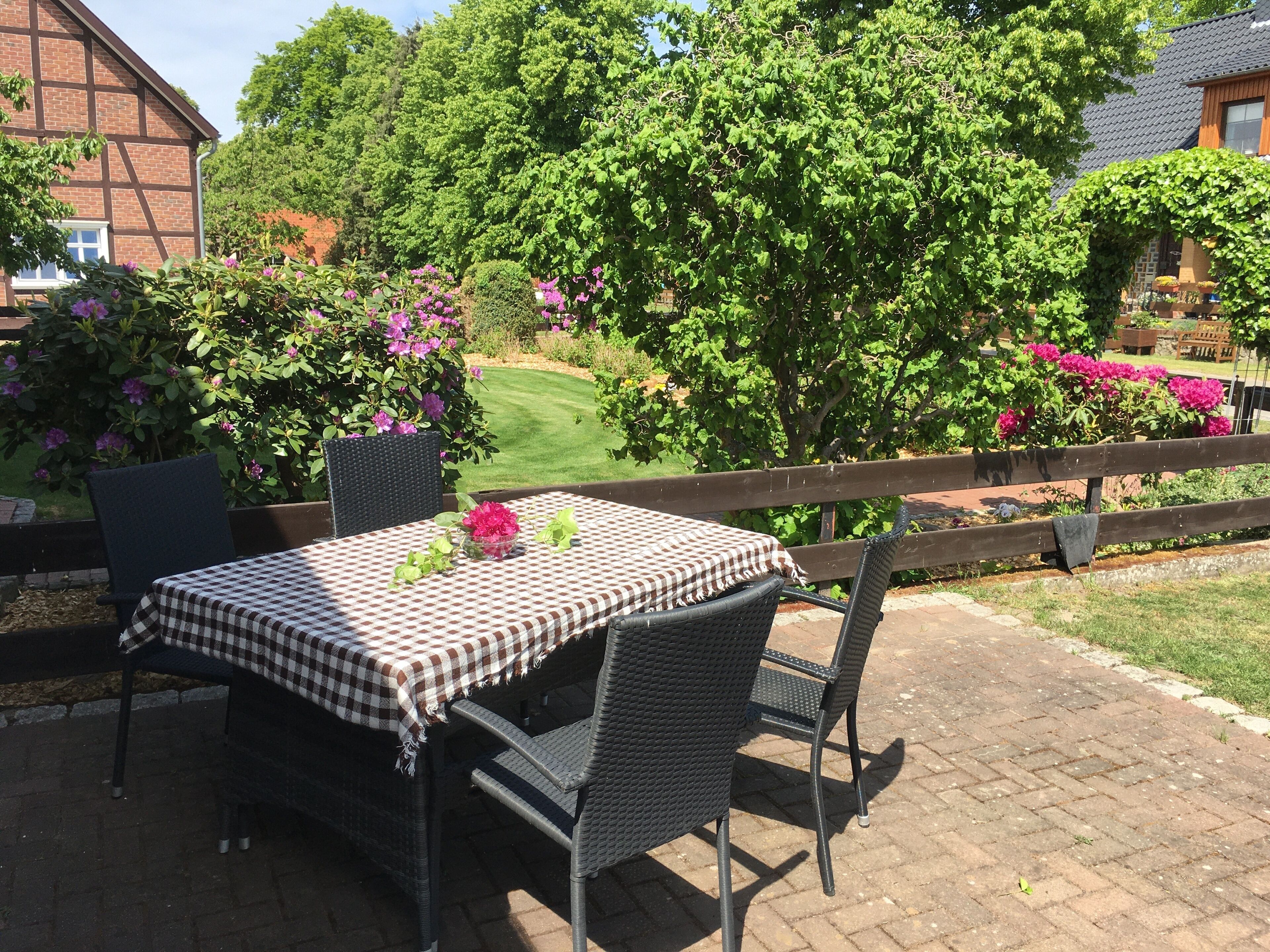 Une terrasse extérieure avec une nappe à carreaux, des chaises en osier et une vue sur un jardin agrémenté d’arbustes en fleurs et d’une pelouse.