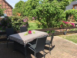 Une terrasse extérieure avec une nappe à carreaux, des chaises en osier et une vue sur un jardin agrémenté d’arbustes en fleurs et d’une pelouse.
