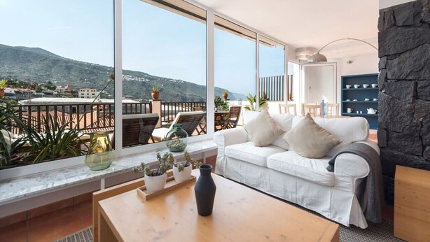 A modern living room with a white sofa, a coffee table, and a view of a balcony with plants and a cityscape in the background. - Ocean/volcano views, terrace, wifi, concierge [B] (Icod de los Vinos)