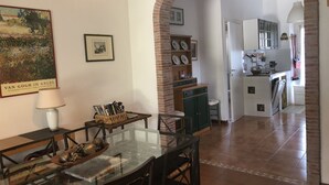 A dining area with a glass table, chairs, and a lamp. There is a brick archway leading to a kitchen with a sink and cabinets.