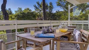 Une table en bois sur laquelle se trouvent un livre, une carafe de jus d’orange et des verres, le tout placé sur un balcon doté de balustrades blanches, avec des arbres en arrière-plan.