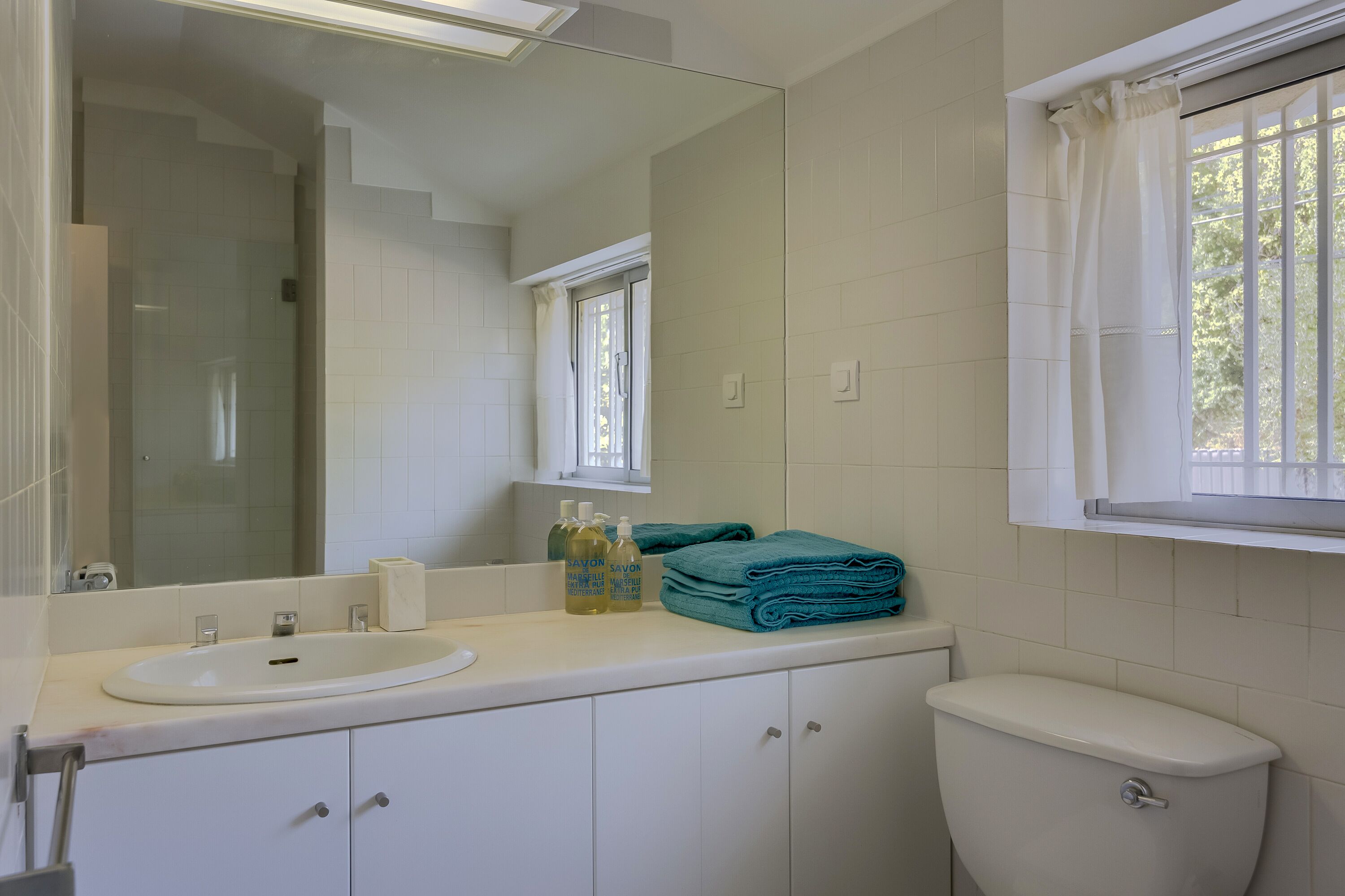 A bathroom with a white sink, mirror, and toilet. There are two bottles of Savon soap and a stack of blue towels on the counter.